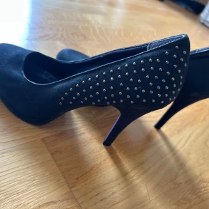 Svarta pumps med nitar från Tamaris - Snygga svarta pumps från Tamaris med mockaliknande material och coola silvriga nitar på hälen. Skorna har hög klack och rund tå, samt lila sula undertill som ger en unik touch. Perfekta för dig som vill sticka ut lite extra.