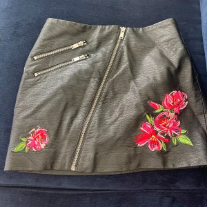 Svart skinnkjol - Cool svart kortkjol i skinnimitation med broderade röda och rosa blommor framtill. Kjolen har två sneda dragkedjor upptill och en längre dragkedja som går diagonalt över framsidan. Perfekt statement-piece med edgy vibe.