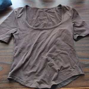 Brun kortärmad topp med vid urringning - En basic brun topp i bomull med korta ärmar och en bred, djup urringning framtill. Modellen är enkel och passar perfekt till jeans eller kjol för en chill look. Tunt och mjukt material som känns skönt mot huden.
