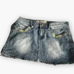 Jeanskjol med broderi från Lonky - Snygg kort jeanskjol från Lonky med slitna detaljer och broderade fickor i blått, gult och orange. Klassisk femficksmodell med rå kant nertill och coola nitar. Perfekt för dig som gillar en edgy och trendig stil.