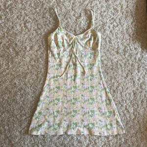 Blommig kortklänning med flätade band - Supersöt kortklänning med blommigt mönster i pastelliga färger som rosa, gul, grön och vit. Klänningen har smala flätade axelband, knytning fram och en öppen rygg med snygga korsade band. Perfekt för varma dagar och ger en riktigt somrig vibe!