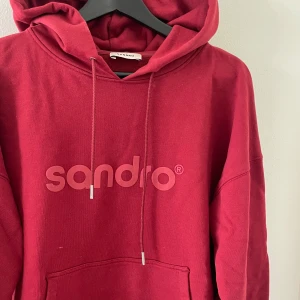 Röd hoodie från Sandro Paris - Snygg röd hoodie från Sandro med stor logga framtill. Tröjan har huva med dragsko och en klassisk känguruficka. Perfekt för dig som gillar en avslappnad och trendig stil. Tillverkad i mjuk bomull för extra komfort. Nypris: 2999kr