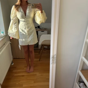 Ljusgul kort klänning med broderi - Superfin adoore ljusgul kort klänning med v-ringning, markerad midja och stora puffärmar. Klänningen har knappar framtill och är täckt av broderade blommor som ger en romantisk känsla. Perfekt för sommarens alla tillfällen!