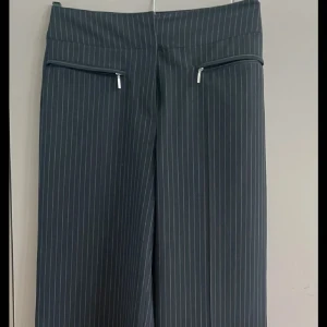 Svarta randiga kostymbyxor med dragkedjor - Snygga svarta kostymbyxor med smala vita ränder och två dragkedjor framtill. Byxorna har hög midja och vida ben för en trendig look. Perfekta till en cool oversized skjorta eller stickad tröja. Materialet känns mjukt och följsamt.