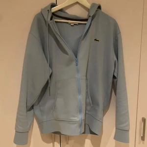 Ljusblå hoodie från Lacoste - Säljer en ljusblå hoodie från Lacoste med dragkedja och klassisk krokodillogga på bröstet. Tröjan har huva med snörning, känguruficka och långa ärmar. Perfekt för en chill och avslappnad stil.