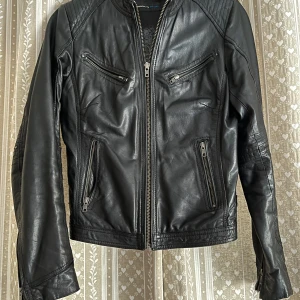 Svart skinnjacka från Rock'n Blue - Säljer en svart skinnjacka från Rock'n Blue med coola detaljer som dragkedjor på bröstet och ärmarna samt quiltade axelpartier. Jackan har figursydd passform och snygga spännen i midjan. Fodret har ett unikt tryck som ger extra edge.