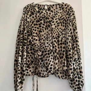 Leopardmönstrad blus med snörning - Säljer en snygg blus i leopardmönster med v-ringning och långa ärmar. Toppen har en cool snörningsdetalj på sidan och är gjord i ett glansigt, lätt material som känns lyxigt. Perfekt för dig som vill sticka ut med djurmönster och trendig look.