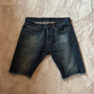 Slim jeansshorts från Ralph Lauren - Snygga jeansshorts från Ralph Lauren Denim & Supply med klassisk femficksdesign och råa, fransiga benslut. Mörkblå tvätt med slitningar och cool patch med amerikanska flaggan bak. Perfekt för dig som gillar street och avslappnad stil.