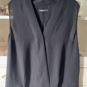 Svart blus från Gina Tricot - Stilren svart blus från Gina Tricot i storlek 40. Västen har en omlott-design med djup v-ringning och är ärmlös. Materialet är mjukt och lätt, perfekt för att bygga lager eller ge en clean look. Passformen är loose och blusen är helt enfärgad utan detaljer.
