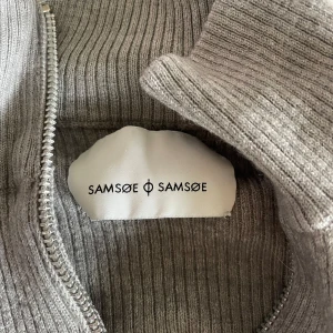 Grå ribbad half zip tröja Samsøe Samsøe - Säljer en grå ribbad långärmad tröja från Samsøe Samsøe med half zip och hög krage. Tröjan är stickad i mjukt material och har en clean, slim passform som är perfekt för lager-på-lager. Enkel att styla med jeans eller cargo pants.