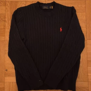 stickad tröja Ralph Lauren - Kabelstickad blå tröja från Polo Ralph Lauren i storlek L. Klassisk modell med rund hals och röd broderad logga på bröstet.  Liten i storlek så passar bättre M.  Utöver det fint skick.
