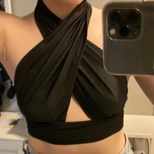Svart omlott halterneck topp - Säljer en svart omlott topp med halterneck och croppad passform. Toppen har en snygg twistad design framtill och är ärmlös, perfekt för att skapa en cool och trendig look. Tillverkad i mjukt och stretchigt material som sitter skönt mot kroppen.