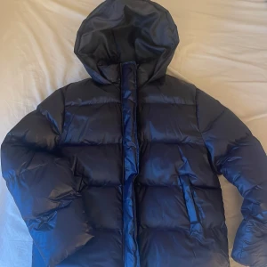 Svart pufferjacka med huva - Säljer en svart pufferjacka med stor huva och breda, quiltade sektioner. Jackan har dragkedja framtill och två snedställda fickor. Perfekt för kalla dagar när du vill hålla värmen och ändå se chill ut. Oanvänd så skicket är toppen. Storleken är S men den passar mer en M.