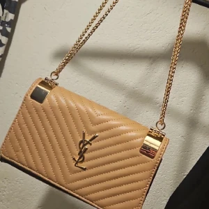 Beige quiltad YSL axelväska i skinn - Säljer en beige axelväska från YSL med klassisk quiltad design och guldfärgade detaljer. Väskan har kedjerem i guld och ikoniska YSL-loggan framtill. Tillverkad i skinn och perfekt för dig som vill ha en stilren och lyxig accessoar.