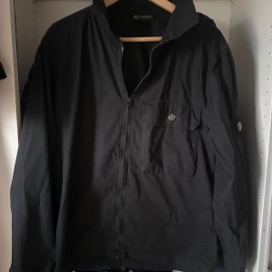 Svart vindjacka från Lagency, M - Svart vindjacka från Lagency i storlek M. Jackan har dragkedja framtill, bröstficka med knapp och detaljerade knappar på ärmarna. Lätt och smidig modell med klassisk krage, perfekt för dig som gillar stilren och enkel design. Defekt på  vänster arm bestående av två hål och lite skrap defekter. 