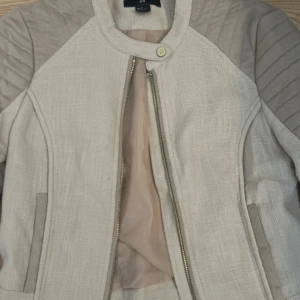 Beige bomberjacka från H&M, stl 38 - Snygg beige bomberjacka från med detaljer på axlar och ärmar. Den har dragkedja, rund krage och två fickor. Jackan är tajt i ärmarna och kan kännas mindre i storleken. Materialet är en mix av bomull och syntet, vilket ger en stilren och modern look.