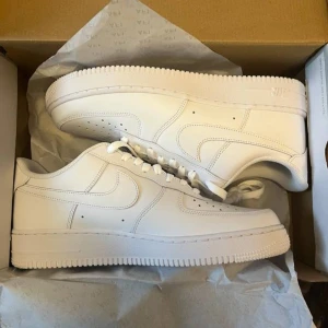 Nike Air Force 1 helvita sneakers - Klassiska Nike Air Force 1 sneakers i helvitt skinn med låg profil och rund tå. Snygg och clean design med perforerade detaljer på tån och ikoniska swoosh-loggan på sidorna. Perfekta för dig som gillar stilrena och tidlösa sneakers.