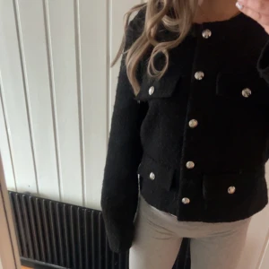 Svart cardigan jacka från H&M, M - Populär Stilren svart cardigan kofta från H&M i medium. Koftan har grovstickad känsla, fyra fickor framtill och coola silvriga knappar. Rak passform och långärmad modell, perfekt för att lyfta vilken outfit som helst. (Den är sydd på högra sida, syns på 5 bilden)