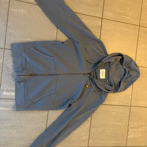 Blå hoodie från Lyle & Scott - Säljer en blå hoodie från Lyle & Scott med dragkedja och huva. Tröjan har två stora fickor framtill och den klassiska gula loggan på bröstet. Perfekt för chill dagar eller när du vill ha en enkel och snygg look.