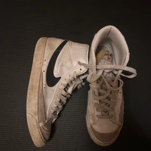 Nike Blazer Mid vit/svart sneakers - Nike Blazer Mid sneakers i vitt skinn med svart swoosh på sidan och tydlig Nike-logga på hälen. Klassisk hög modell med snörning och platt sula. Insidan är vadderad och skon har en rund tå. Perfekt för dig som gillar retrostil och streetwear.