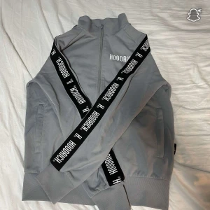 Grå track jacket från Hoodrich - Snygg grå track jacket från Hoodrich med svart band längs ärmarna där det står HOODRICH i vitt. Jackan har hög krage, dragkedja framtill och ribbade muddar. Perfekt för en sportig och street-inspirerad look.
