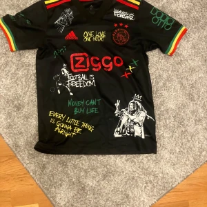 Ajax x Bob Marley Adidas fotbollströja S - Unik svart Ajax fotbollströja från Adidas med Bob Marley-tema och färgglada detaljer i rött, gult och grönt. Tryck med texter, illustrationer och klubbmärke på bröstet. Tillverkad i lätt och ventilerande Aeroready-material. Perfekt för dig som vill sticka ut på läktaren eller planen.