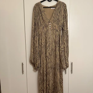 Zebramönstrad långklänning från Adoore - Snygg långklänning från Adoore med zebramönster i beige och brunt. Klänningen har v-ringning, knappar framtill och långa, puffiga ärmar. Perfekt för dig som vill sticka ut med ett coolt mönster och avslappnad passform.