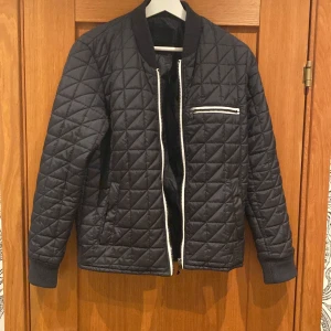 Svart quiltad bomberjacka XS - Quiltad svart bomberjacka från SS 1995 i storlek XS. Jackan har vita dragkedjor, ribbade muddar och en bröstficka med dragkedja. Snyggt rutmönster över hela jackan och klassisk bomberkrage. Perfekt för dig som gillar stilren look!