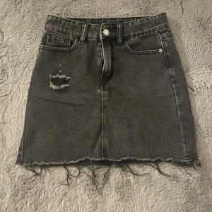 Svart jeanskjol med slitna detaljer - Cool svart jeanskjol med rå kant nertill och slitning framtill. Kjolen har klassiska fickor både fram och bak samt bälteshällor. Perfekt för dig som gillar en edgy och avslappnad stil.