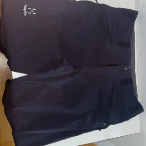 Svarta täckbyxor från Haglöfs - Säljer ett par svarta täckbyxor från Haglöfs med dragkedjor på fickorna och justerbara detaljer i midjan. Perfekta för kalla dagar och utomhusaktiviteter. Snygg och funktionell design med hög komfort.