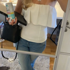 Vit axelbandslös volangtubtopp - Säljer en superfin vit tubtopp med volangdetalj upptill. Toppen är axelbandslös och har en bandeauform som sitter snyggt och markerar midjan. Perfekt till jeans eller kjol för en fräsch och trendig look.