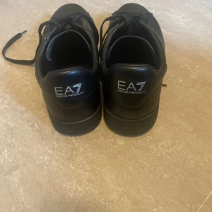 Svarta EA7 Emporio Armani sneakers - Säljer ett par svarta EA7 Emporio Armani sneakers i skinn med klassisk logga på sidan och hälen. Skorna har snörning och en platt sula, perfekt för dig som gillar stilrena och snygga sneakers med premiumkänsla.