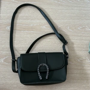 Mörkgrön  axelväska med metallspänne - Stilren mörkgrön axelväska i skinnimitation med justerbart axelband och coolt metallspänne framtill som har ormmönstrad detalj. Väskan har två fack inuti och stängs med magnetlås. Perfekt storlek för det viktigaste och ger en edgy vibe till din outfit.