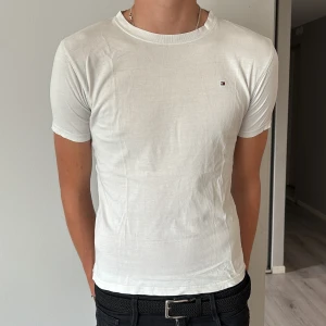 Vit t-shirt från Tommy Hilfiger - Vit t-shirt från Tommy Hilfiger med klassisk rund hals och liten broderad logga på bröstet. Materialet är mjuk bomull som sitter skönt mot kroppen. Perfekt för dig som gillar clean och stilren look.