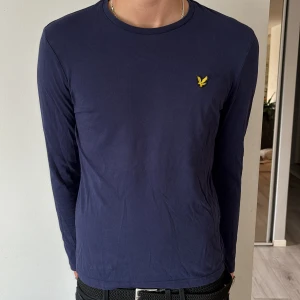 Marinblå långärmad tröja Lyle & Scott - Säljer en marinblå långärmad tröja från Lyle & Scott med klassisk rund hals och gul broderad logga på bröstet. Tröjan är i mjuk bomull och har en clean, stilren look som funkar till allt. Perfekt för dig som gillar enkel och snygg stil.