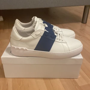 Valentino Garavani vita sneakers med blå detalj - Snygga vita sneakers från Valentino Garavani med bred blå rem över vristen och ikoniska nitar på hälen. Skorna har rund tå, platt sula och är tillverkade i skinn. Perfekta för dig som vill ha en clean men ändå unik look. Skorna är storlek 46 men passar även 45 alldeles perfekt. Alltså lite små i storleken. Hör av er vid ev frågor. Pris går att diskutera vid snabb affär