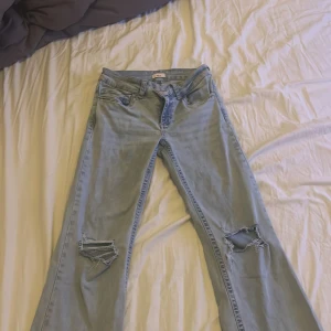Replay ljusblå bootcut jeans med slitningar - Säljer ett par ljusblå Replay jeans med bootcut passform och låg midja. Jeansen har slitna detaljer vid knäna och klassiska röda Replay-broderier på bakfickorna. Tillverkade i mjukt denim-material som sitter snyggt och bekvämt.