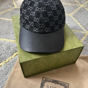 Svart keps från Gucci med GG-mönster - Snygg svart keps från Gucci med klassiskt GG-monogram i grått över hela ovandelen. Skärmen är i svart skinn och resten av kepsen är i jeansmaterial. Perfekt för dig som vill ha en lyxig och trendig accessoar med tydlig designerstil. Storlek M Nya modellen. Möts upp i Göteborg 