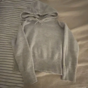 Ljusgrå stickad hoodie med huva - Mysig ljusgrå stickad hoodie med huva och snörning framtill. Tröjan har långa ärmar och ribbade muddar vid ärmslut och nederkant. Perfekt för kyliga dagar när du vill vara både bekväm och stilren.