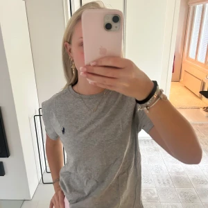 Grå t-shirt från Ralph Lauren - Klassisk grå t-shirt från Ralph Lauren med mörkblå broderad logga på bröstet. Modellen är rak med rund halsringning och korta ärmar. Skön och enkel stil som funkar till det mesta. Materialet är mjuk bomull.