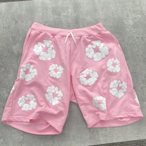 Rosa shorts från Denim Tears med blommor - Säljer ett par rosa shorts från Denim Tears med vita blommiga tryck över hela plagget. De har resår i midjan med snörning och är tillverkade i mjuk bomull, perfekta för en chill sommarlook.