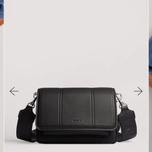 Svart axelväska från NA-KD i skinn - Snygg svart axelväska från NA-KD i skinnimitation med bred justerbar axelrem och stilrena sömmar. Väskan har ett lock med diskret logga framtill och silverfärgade metalldetaljer. Perfekt för att bära dina essentials med stil.