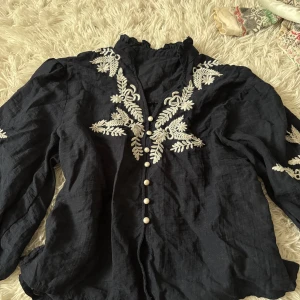 Svart blus med broderi och pärlknappar - Supersnygg svart blus med vita broderade detaljer framtill och på ärmarna. Hög krage med volang, långa puffärmar och breda smockade muddar. Stängs med dekorativa pärlknappar. Perfekt för dig som gillar unika detaljer och vintagevibbar. Ny endast testad