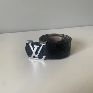 Louis Vuitton svart rutigt skärp - Snyggt svart skärp från Louis Vuitton med klassiskt rutigt mönster och stor LV-logga i silver på spännet. Bältet är gjort i skinn och har exklusiva detaljer samt präglad logga på insidan. Perfekt accessoar för att lyfta din outfit.