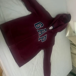 Vinröd hoodie från GAP Kids - Mysig vinröd hoodie från GAP Kids med stor logga framtill i vitt och mörkblått. Tröjan har huva, dragkedja och två fickor framtill. Perfekt för chill dagar och enkel att matcha med jeans eller mjukisbyxor.
