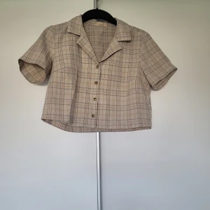 Beige rutig croppad skjorta Urban Renewal - Kortärmad croppad skjorta från Urban Renewal i beige med rutigt mönster. Skjortan har klassisk krage, knappar framtill och en avslappnad passform. 