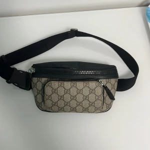 Äkta Gucci beige väska - Säljer en snygg magväska från Gucci i beige canvas med klassiskt GG-mönster och svarta skinn- och textildetaljer. Väskan har flera fack med dragkedja och justerbart band. Perfekt för att bära det viktigaste på ett stilrent sätt. Den är såklart äkta, fråga gärna frågor så svarar jag nästan direkt! Serie nummer och äktighetsbevis finns!