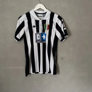 Retro Juventus fotbollströja - Juventus fotbollströja med klassiska svarta och vita ränder, korta ärmar och krage. På framsidan finns klubbmärke, sponsortryck och Kappa-logga. Baksidan har röd text med Zidane och nummer 21. Materialet är lätt och andas, perfekt för match eller träning.