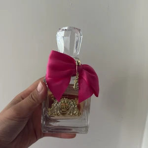 Juicy Couture parfym med rosa rosett - Juicy Couture parfym i en lyxig, fyrkantig glasflaska med en stor, kristalliknande kork. Flaskan är dekorerad med en starkt rosa rosett och guldfärgad emblem med krona framtill. Perfekt för dig som gillar söta och feminina dofter.