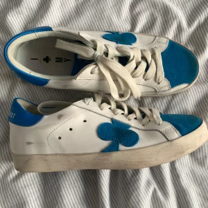 Vita och blå sneakers med klubbmärke - Unika vita sneakers med blå detaljer och klubbmärke på sidan. Skorna har blå mockadetaljer på tå och häl, samt snörning framtill. Tillverkade i skinn och mocka med platt sula och rund tå. Perfekta för dig som vill sticka ut med en cool och lekfull stil.
